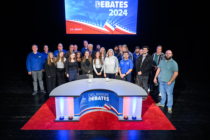 A group of men and women gathered inside the Delaware Debates studio, standing behind the red white and blue desk and in front of a sign with the Delaware Debates logo.